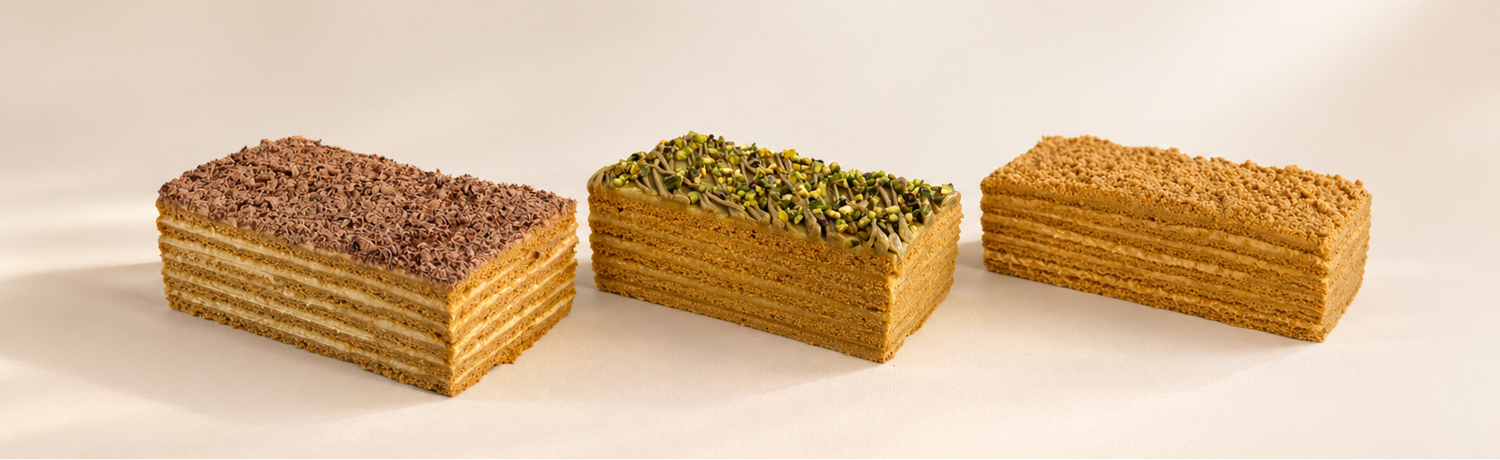 Three layered Medovik honey cakes with chocolate, pistachio, and classic toppings on a neutral background by Minella Patisserie Melbourne
